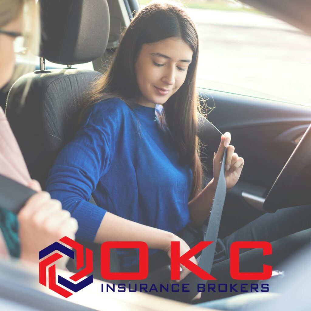 A teenage girl buckles her seatbelt in the driver’s seat while her mom sits beside her, watching and coaching during a driving lesson.